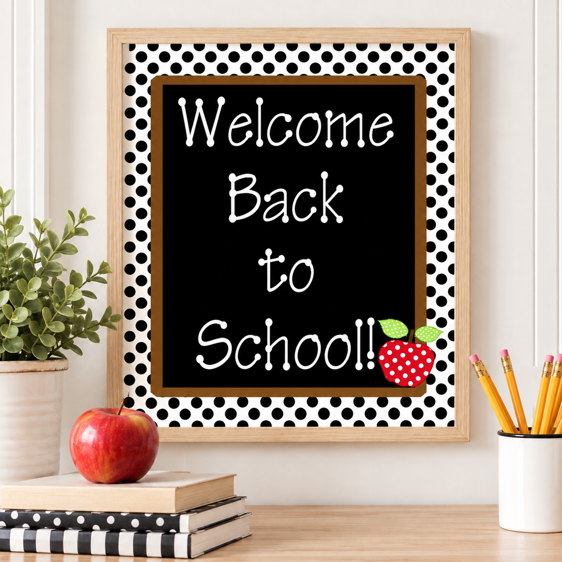 A framed 'Welcome Back to School' sign with a black center, white polka dot border, and a red apple illustration, displayed on a desk with books, an apple, and pencils.