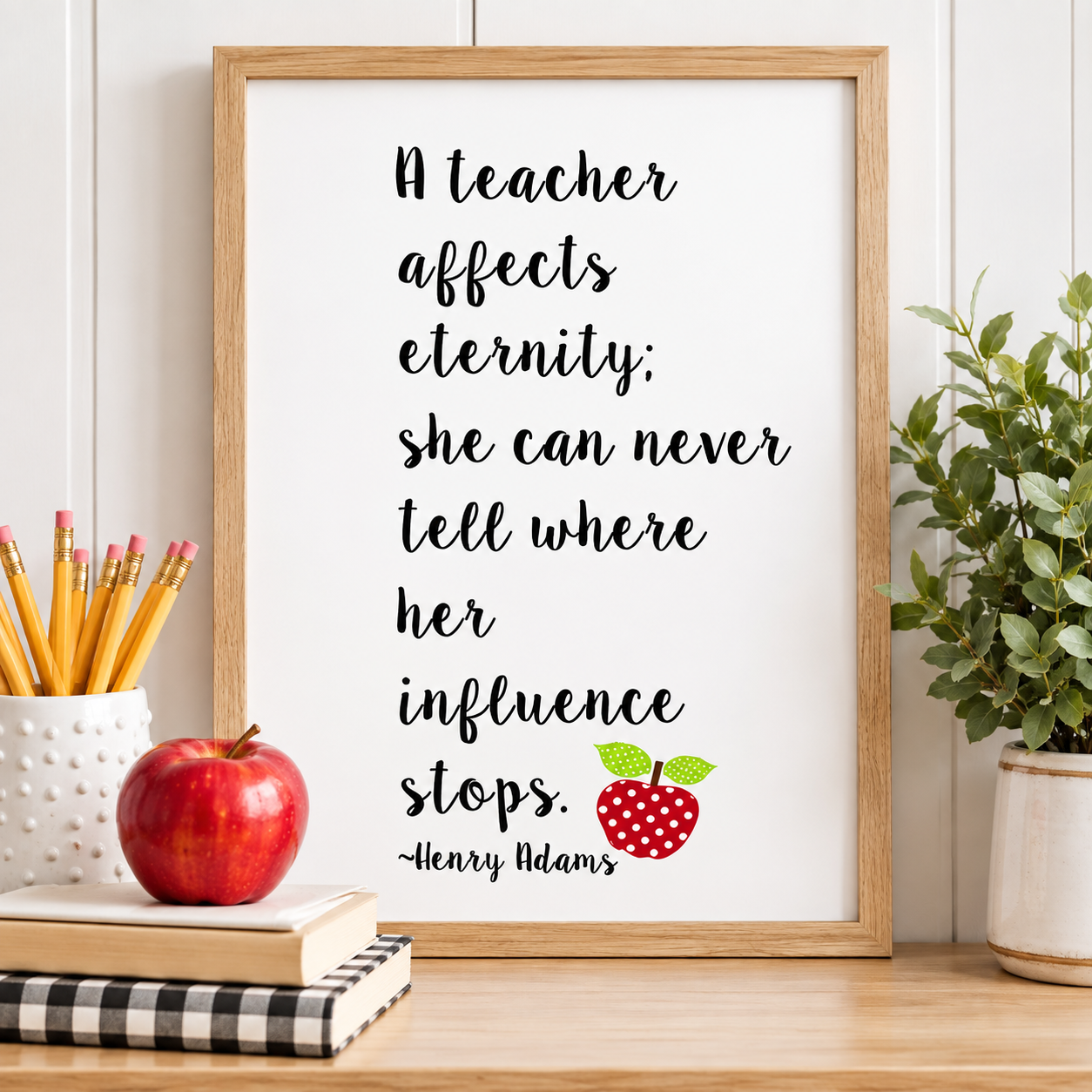 A framed typographic wall art print for a teacher with a black quote on a white background, styled on a wooden shelf with a red apple, pencils, a notebook, and a potted plant. Teacher appreciation gift.
