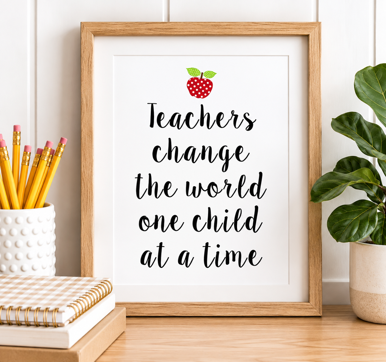 A framed typographic wall art print with the message “Teachers change the world one child at a time” in black script, topped with a small red apple illustration. The print is shown in a light wood frame on a tabletop, with pencils in a white cup, stacked notebooks, and a green plant nearby.