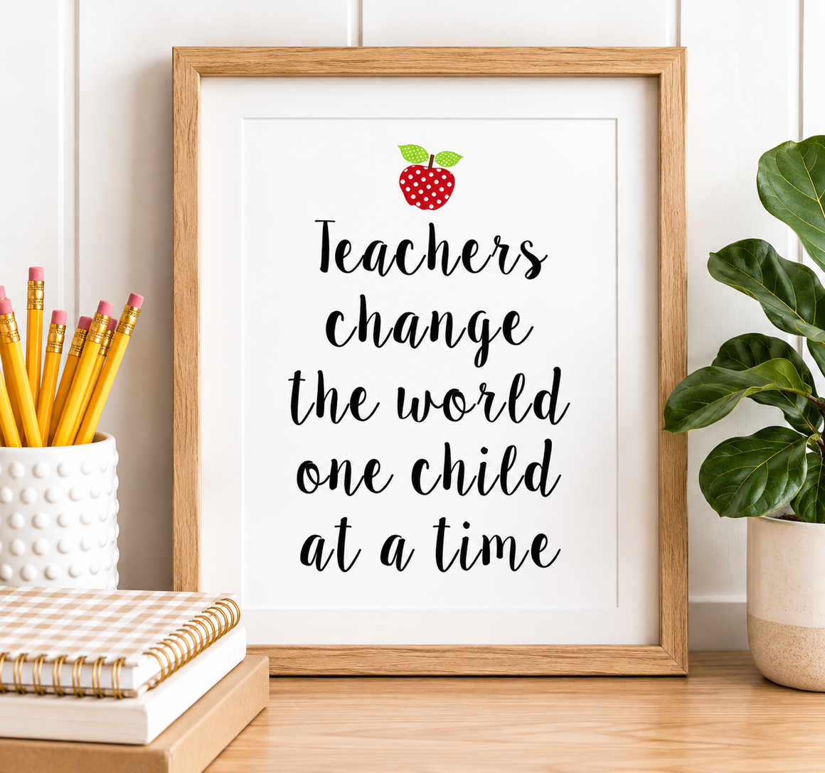 A framed typographic wall art print with the message “Teachers change the world one child at a time” in black script, topped with a small red apple illustration. The print is shown in a light wood frame on a tabletop, with pencils in a white cup, stacked notebooks, and a green plant nearby.