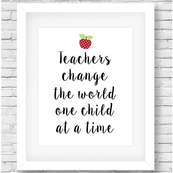 A framed typographic wall art print with the message “Teachers change the world one child at a time” in black script, topped with a small red apple illustration. The print is shown in a light wood frame on a tabletop, with pencils in a white cup, stacked notebooks, and a green plant nearby.